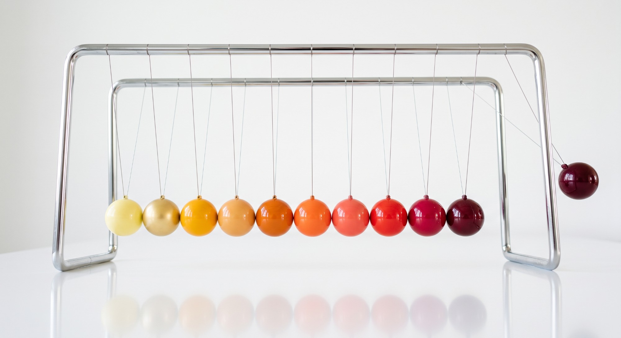 A Newton's cradle of ten polished spheres in a warm gradient from pale yellow to deep burgundy, the end ball raised and about to strike